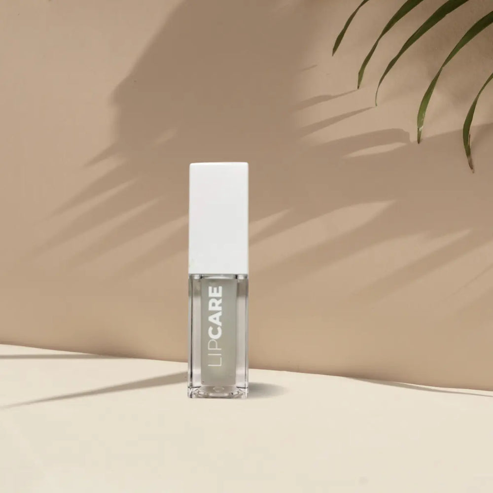 En närbild av en vit flaska med en transparent etikett där texten "Lipcare" är synlig. Flaskan är fylld med en klar eller vitaktig produkt. Bakgrunden är beige/brun. Produkten är designad för att ge maximal fukt och vård till läpparna och är vegansk och cruelty-free.