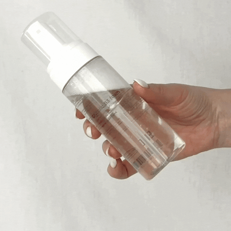 Vit flaska med svart text, visad i animerad rörelse mot ljus bakgrund – ett exfolierande rengöringsskum för alla hudtyper.