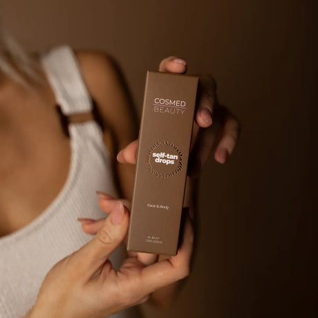 En brun pappkartong med produktnamn och beskrivning tryckt tydligt på framsidan. Kartongen innehåller produkten "Self Tan Drops" från Cosmed Beauty, designad för att ge en naturlig, solkysst lyster genom att blandas med din dagliga fuktighetskräm – helt utan skadliga UV-strålar.
